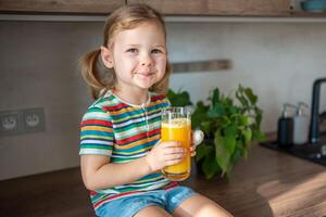 poco ragazza potabile fresco succo seduta su tavolo nel casa cucina foto