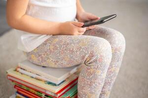 poco ragazza si siede su un' pila di figli di libri e usi smartphone foto