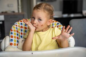 poco ragazza con biondo capelli mangiare fatti in casa cioccolato e mostrando bocca e sporco mani con macchie di cioccolato nel casa cucina foto