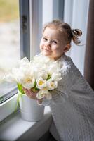 poco ragazza seduta di finestra con tulipano fiori mazzo. contento bambino, al chiuso. La madre di giorno, San Valentino giorno o compleanno concetto. foto