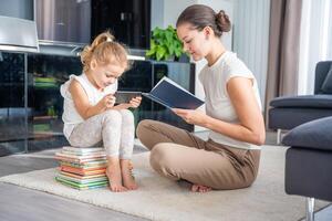poco ragazza si siede su un' pila di figli di libri e usi sua smartphone mentre madre legge un' libro foto