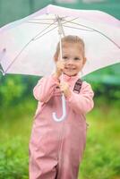 contento ridendo bambino ragazza 2-3 anno vecchio indossare impermeabile Abiti e Tenere rosa ombrello avere un' divertimento su casa Giardino dietro la casa nel piovoso giorno foto