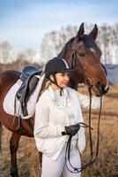 bella biondo professionale femmina fantino in piedi vicino Marrone cavallo nel campo. amicizia con cavallo. alto qualità foto