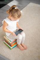 poco ragazza si siede su un' pila di figli di fiaba libri e orologi cartoni animati su sua smartphone foto