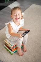 sorridente poco ragazza si siede su un' pila di figli di fiaba libri e orologi cartoni animati su sua smartphone foto