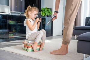 poco ragazza si siede su un' pila di figli di libri e usi sua smartphone mentre madre dà sua un' libro foto