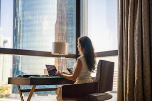 elegante donna d'affari libero professionista Lavorando con computer portatile, lei è seduta su il sedia, godendo panoramico Visualizza nel finestra con città sfondo. Basso chiave foto