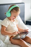 poco ragazza nel cuffie utilizzando digitale tavoletta e sorridente contento mentre ascoltando per musica o giocando gioco nel casa letto. alto qualità foto