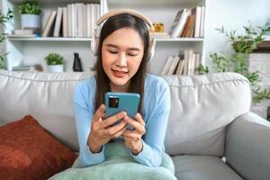 felicemente rilassato giovane donna si siede su un' divano utilizzando un' mobile Telefono, ascoltando per musica attraverso un App e cuffie foto