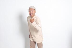 ritratto di scioccato asiatico musulmano uomo nel koko camicia con zucchetto mostrando Prodotto e puntamento con pollice su per il lato. pubblicità concetto. isolato Immagine su bianca sfondo foto
