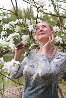 giovane biondo donna con lungo capelli sta nel il parco Il prossimo per un' fioritura Mela albero, primavera tempo, unità con natura, foto