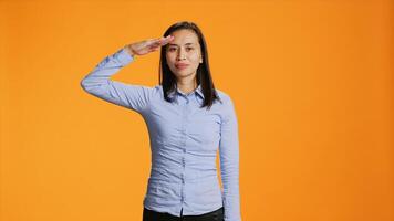 patriottico donna mostrando militare saluto gesto su telecamera, raccolta mano al di sopra di templi e in piedi orgoglioso nel studio. asiatico persona salutando con esercito simbolo contro arancia sfondo. foto