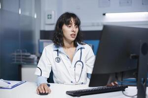 focalizzata femmina medico utilizzando un' del desktop pc nel un' ospedale ufficio. nel un' clinica camera, un' caucasico medico indossare un' laboratorio cappotto, utilizzando un' computer per dai un'occhiata medico File. foto