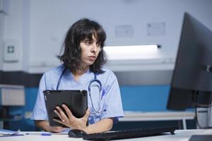 donna Lavorando come un' infermiera con digitale tavoletta e computer per medico informazione e verifica. assistenza sanitaria specialista Tenere un' dispositivo con toccare schermo mentre controllo paziente dati su del desktop pc. foto