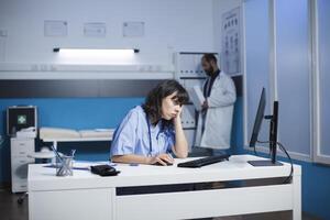 dopo difficile giorno a Ospedale, un' stanco caucasico infermiera seduta a un' scrivania con un' del desktop computer. femmina professionista carezzevole sua viso a un' clinica ufficio, guardare esausto. foto