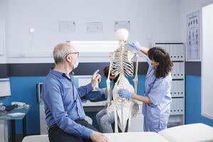 anziano paziente guardare a umano scheletro per ortopedico assistenza sanitaria visita medica. femmina infermiera professionista puntamento a spinale cordone, spiegando diagnosi per osteopatia e recupero per anziano uomo. foto