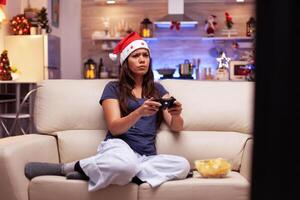 ragazza gamer giocando in linea videogiochi utilizzando gioco telecomando da gioco durante virtuale concorrenza nel natale decorato cucina. donna festeggiare inverno stagione godendo Natale vacanza foto