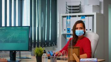 donna con protezione maschera Lavorando e guardare a telecamera seduta nel davanti di pc. dipendenti nel spazio di lavoro nel aziendale azienda digitando su computer tastiera guardare a del desktop rispetto sociale distanziamento foto