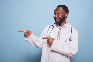 ritratto di allegro maschio medico vestito nel laboratorio cappotto e stetoscopio puntamento Il prossimo per lui su isolato blu sfondo. sorridente africano americano medico Esprimere a gesti con indice dito per il suo giusto lato. foto