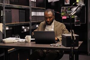 confuso africano americano investigatore ricerca prova utilizzando il computer portatile. preoccupato legge rinforzo professionale seduta a posto di lavoro scrivania e analizzando crimine Astuccio su computer nel sera foto