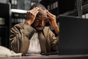 stanco poliziotto sofferenza a partire dal emicrania Lavorando col tempo su il computer portatile. stressato africano americano polizia investigatore con mal di testa soluzione crimine e analizzando rapporti nel ufficio in ritardo foto