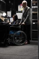 paralizzato asiatico donna d'affari esecutivo seduta nel sedia a rotelle a ufficio scrivania, ispezionando trimestrale risultati analisi dati su il computer portatile. donna con paraplegia nel contabilità posto di lavoro foto