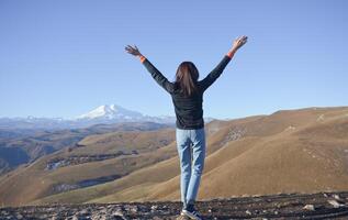 un' giovane brunetta donna con sua mani sollevato su contro il fondale di il snow-capped montare elbrus. foto