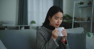 ritratto di rilassante giovane donna Tenere tazza godere odore caffè o tè e potabile con felicità nel mattina mentre sedersi su divano nel vivente stanza, libera tempo, prendi rompere nel a casa, sorridente foto