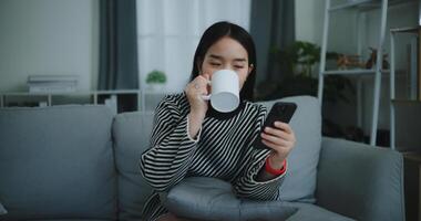 ritratto di contento giovane asiatico donna sedersi su divano godere chat su mobile Telefono mentre potabile caffè nel mattina nel vivente stanza, libera tempo, prendi pausa, sorridendo foto