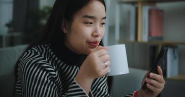 ritratto di contento giovane asiatico donna sedersi su divano godere chat su mobile Telefono mentre potabile caffè nel mattina nel vivente stanza, libera tempo, prendi pausa, sorridendo foto