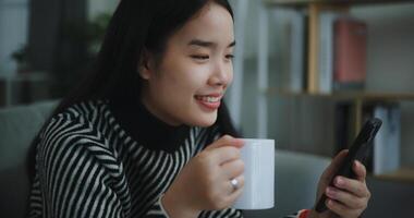 ritratto di contento giovane asiatico donna sedersi su divano godere chat su mobile Telefono mentre potabile caffè nel mattina nel vivente stanza, libera tempo, prendi pausa, sorridendo foto