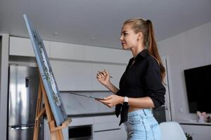 un' creativo femmina artista sta e lavori su un' la pittura. un' contento giovane signora è pittura su tela nel sua appartamento. il concetto di un' giovane La madre di tempo libero foto