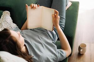 tempo libero volta. giovane donna godendo prenotare, pasto, e tè su vivente camera divano. rilassato lettura e cenare nel casa ambientazione. accogliente stile di vita con libri, cibo, e bevanda. rilassamento nel domestico comfort foto