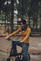 contento giovane asiatico donna mentre equitazione un' bicicletta nel un' città parco. lei sorrise utilizzando il bicicletta di trasporto. l'ambiente amichevole concetto. foto