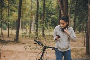 contento asiatico giovane donna camminare e cavalcata bicicletta nel parco, strada città sua sorridente utilizzando bicicletta di trasporto, eco amichevole, persone stile di vita concetto. foto