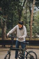 contento asiatico giovane donna camminare e cavalcata bicicletta nel parco, strada città sua sorridente utilizzando bicicletta di trasporto, eco amichevole, persone stile di vita concetto. foto