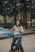 contento asiatico giovane donna camminare e cavalcata bicicletta nel parco, strada città sua sorridente utilizzando bicicletta di trasporto, eco amichevole, persone stile di vita concetto. foto