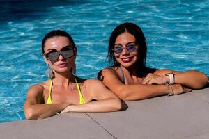 Due attraente ragazze con lungo capelli siamo in posa vicino piscina su il sole. foto