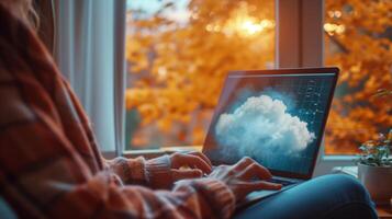 ai generato un' persona lavori su un' il computer portatile con un' a tema nuvola schermo, seduta comodamente a un' casa stazione di lavoro di un' finestra con autunno le foglie fuori. foto