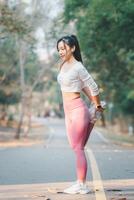 fitness concetto, in forma giovane femmina corridore allungamento sua gamba durante un' riscaldamento nel un' soleggiato parco percorso. foto