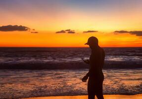 uomo controllo Telefono mentre godendo bellissimo tramonto a piedi su il spiaggia foto