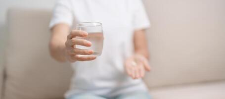 donna mano Tenere medicina antidolorifico pillola e acqua bicchiere su il divano a casa, assunzione per mal di testa, stomaco dolore, diarrea dolore a partire dal cibo avvelenamento, endometriosi, isterectomia e mestruale foto