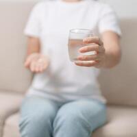 donna mano Tenere medicina antidolorifico pillola e acqua bicchiere su il divano a casa, assunzione per mal di testa, stomaco dolore, diarrea dolore a partire dal cibo avvelenamento, endometriosi, isterectomia e mestruale foto