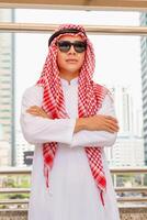 ritratto di arabo uomo d'affari in piedi con braccia attraversato sfocato città sfondo, musulmano uomo nel occhiali da sole in piedi con braccia attraversato foto