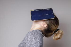 orizzontale Immagine di un' giovane donna copertura sua viso con un' libro. un' bellissimo donna nel un' grigio a maglia maglione, nascondiglio sua viso dietro a un' blu libro coperchio, con sua testa gettato Indietro. libro concetto foto