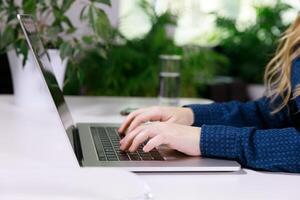 il mani di un invisibile ragazza nel un' blu camicetta siamo digitando su un' il computer portatile a il tavolo. Là è un' mobile Telefono nelle vicinanze e Là siamo molti fiori nel un' pentola. alto qualità foto