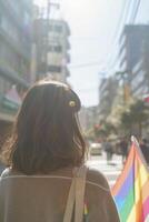 ai generato giovane giapponese donna, indietro trasformato, ornato con un lgbt orgoglio arcobaleno bandiera, in mezzo il pieno di bokeh strade di un' soleggiato giorno orgoglio parata nel il città foto