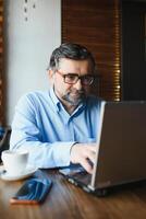 positivo anziano barbuto uomo con grigio capelli potabile caffè e utilizzando il computer portatile a bar, copia spazio. elegante anziano uomo d'affari nel Borgogna giacca godendo il suo tè mentre Lavorando in linea. foto