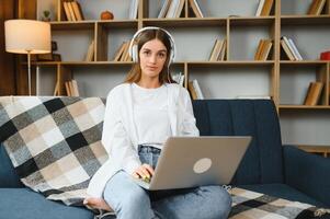 sorridente attraente giovane donna seduta su divano utilizzando il computer portatile comunicare Lavorando in linea a casa, contento adolescente ragazza digitando su computer, godendo scrittura blog o chat con amici nel sociale Rete foto