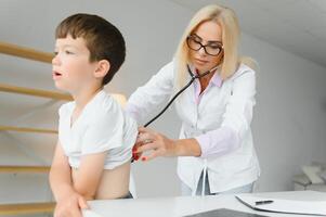 pediatra ascoltando per respirazione nel il polmoni e battito cardiaco con stetoscopio. ritratto di adorabile poco ragazzo visitare medico. foto